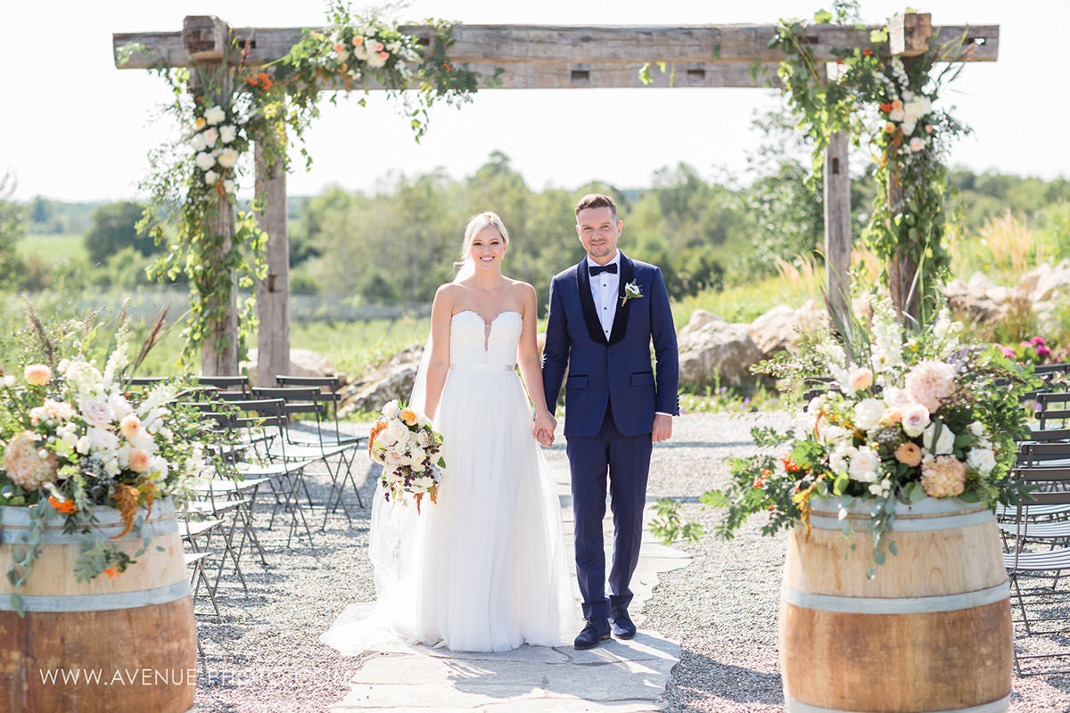 a bride and groom standing together at an outdoor wedding ceremony site overlooking a vineyard under a wooden arbour with florals