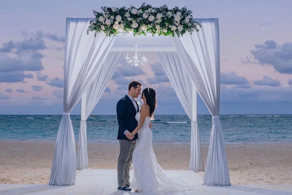 A couple holding hands under a beach-side gazebo with draping and flowers