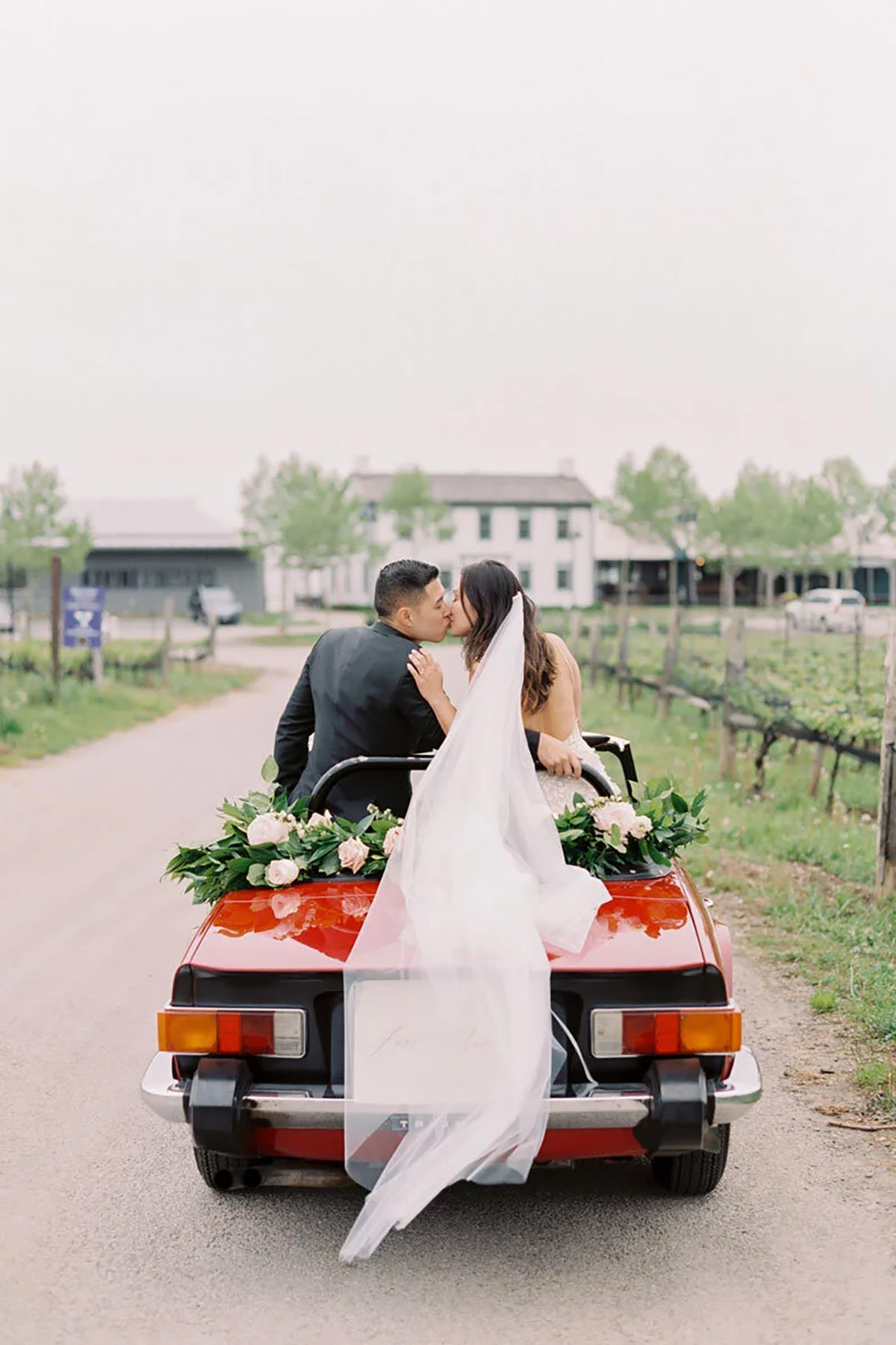 Bride and groom sitting up high in the back seat of a sports car in front of a vineyard