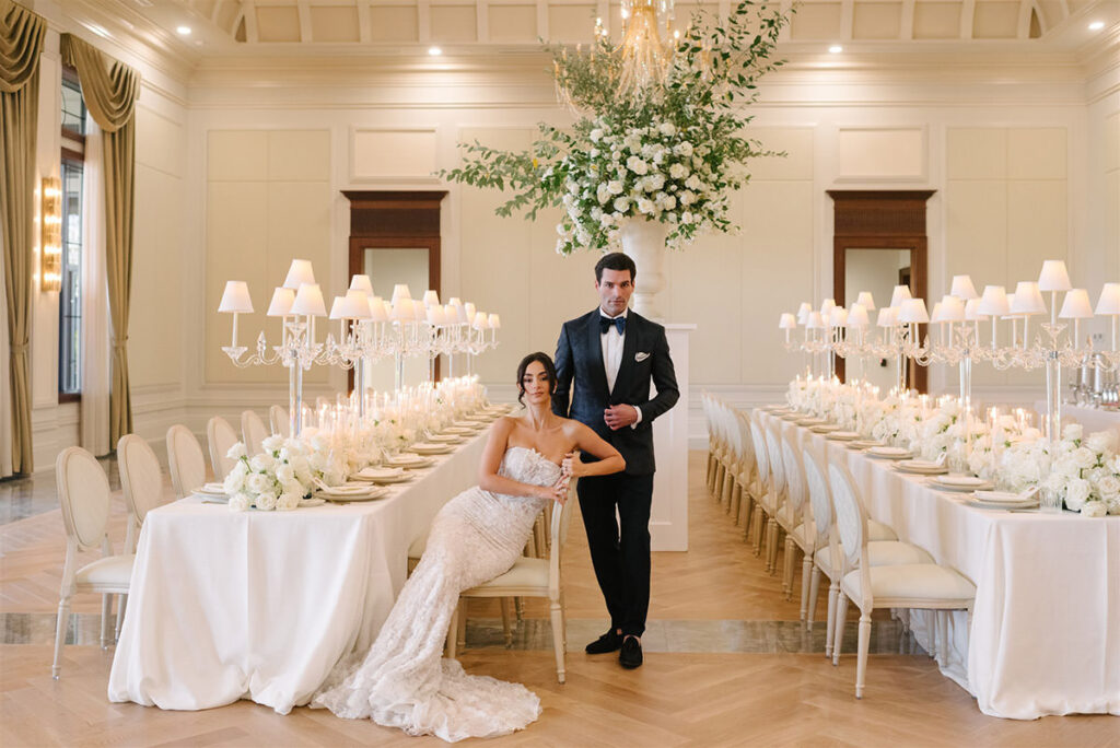 A couple sitting in their reception hall, with white linens and flowers, looking at the camera