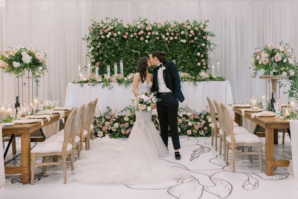 A couple kissing in their reception hall with foliage behind them.