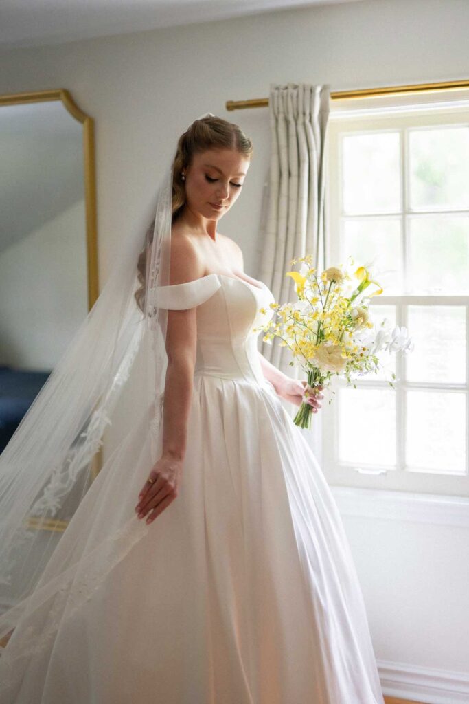 bride in a satin ballgown holding yellow bouquet by a window
