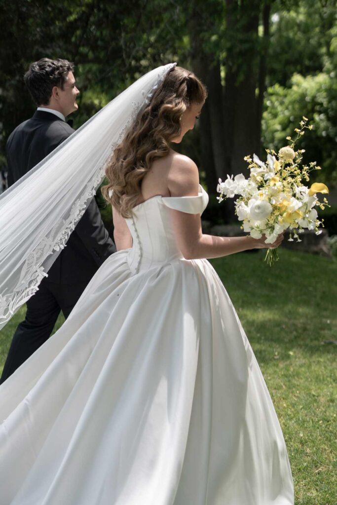 bride in off shoulder satin ballgown and lace veil walking with groom